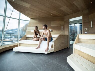 Two people relax in a sauna with large windows and wood interior in a spa. | © TAUERN SPA Zell am See-Kaprun