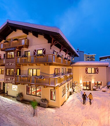 A multi-story hotel in winter snow with lit windows and balcony, surrounded by snowy paths and trees. | © Hotel Glasererhaus