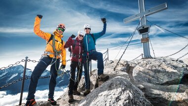 Three mountaineers at the summit of Kitzsteinhorn, standing on rocks next to a cross, with mountain views in the background. | © Kitzsteinhorn 